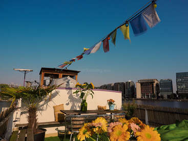 Eine mit bunten Blumen, Pflanzen und Fahnen geschmückte Dachterrasse mit Blick auf einen Fluss und moderne Gebäude unter einem klaren blauen Himmel schafft eine fröhliche, entspannte Atmosphäre.