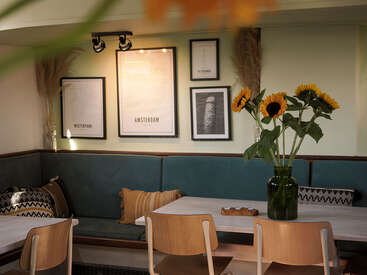 In diesem gemütlichen Café stehen tealfarbene Sitzbänke, moderne Holzstühle und ein weißer Tisch mit Sonnenblumen darauf. Gerahmte, minimalistische Kunst und Kissen schaffen eine einladende Atmosphäre.