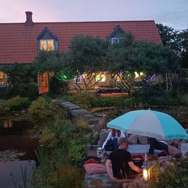 In a cozy garden at dusk, people relax under a blue umbrella with a dog, near a pond, in front of a warmly lit house with trees.