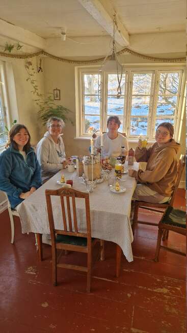 Four people sit around a cozy table by large windows, enjoying breakfast together. Sunlight streams in, snowy landscape outside, creating a warm, inviting atmosphere with rustic decor.