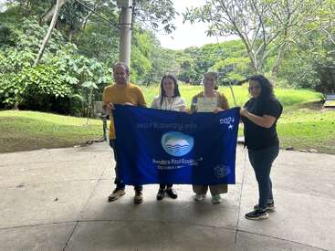 Four people stand outdoors holding a blue "Bandera Azul Ecológica" 2024 flag, smiling. One person holds a certificate. Trees and greenery are in the background.