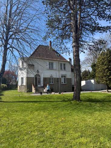 A charming two-story house with a red-tiled roof sits surrounded by trees and green lawn. A person and several dogs are outside enjoying sunshine.