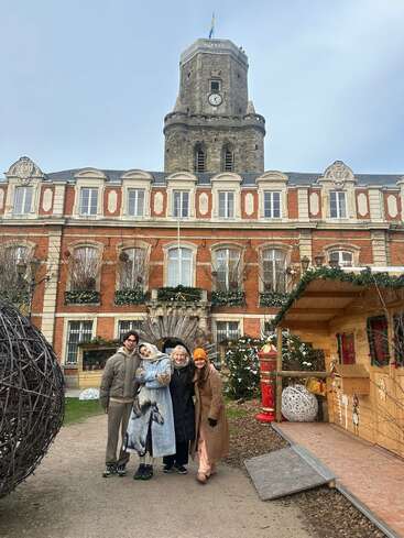 Four people wearing winter clothing pose happily in front of a historic building with a clock tower, festive decorations, and a wooden hut on a cloudy day.