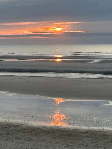 A serene beach at sunset, with orange and yellow hues reflecting on wet sand and shallow pools. Gentle waves meet a calm horizon beneath dramatic clouds.