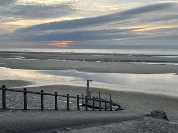 A serene beach scene at sunset, with calm waters reflecting the colorful sky, clouds stretching across, and a wooden railing leading down to the sandy shore.