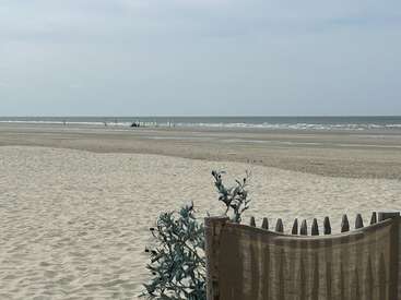 A quiet beach scene with gentle waves meeting sandy shore. Sparse vegetation and wooden fence in the foreground. Few people and sails visible in the distance.