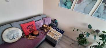 Ein gemütlicher Raum mit einer grauen Couch, bunten Kissen und Musikinstrumenten wie Trommeln und Klangschalen. Kunst hängt an der Wand und Pflanzen schmücken die Ecke.