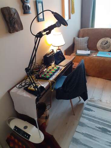 Ein gemütliches Home-Recording-Setup mit einem Mikrofon, einer Loop-Station, einem Laptop, einer Lampe und dekorierten Wänden. Couch, Kissen, Trommel und Papiere sorgen für eine entspannte, kreative Atmosphäre.
