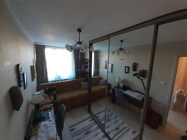 Dieses Bild zeigt ein gemütliches, helles Zimmer mit einem braunen Sofa, einem verspiegelten Kleiderschrank, einem Schreibtisch, Lampen, Dekorationen an den Wänden, einem Teppich und einem Fenster mit Vorhängen.