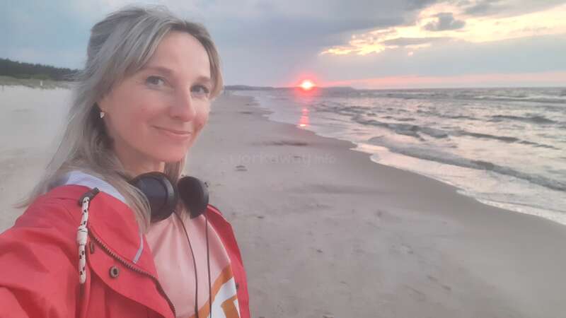 Eine Frau lächelt, während sie ein Selfie an einem Sandstrand bei Sonnenuntergang macht. Sie trägt eine rote Jacke und Kopfhörer. Hinter ihr sind Wellen und dramatische Wolken zu sehen.