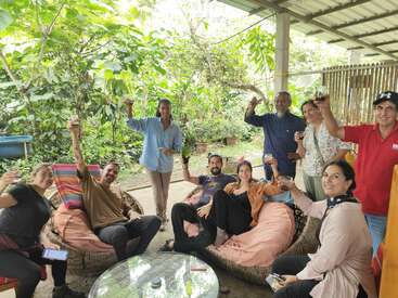 Un grupo de personas, sentadas y de pie, se encuentra en un patio cubierto, sonriendo y levantando las copas en un alegre brindis, rodeadas de exuberantes plantas verdes.