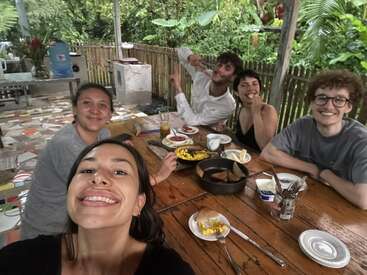 Cinco amigos están sentados alrededor de una mesa rústica de madera al aire libre, sonriendo y disfrutando juntos del desayuno, rodeados de una exuberante vegetación y de un ambiente tropical acogedor y agradable.