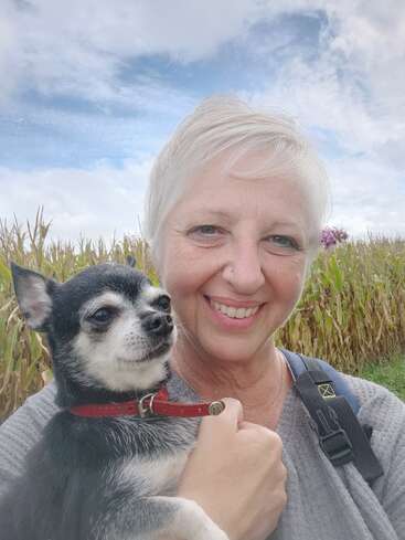 Eine lächelnde Frau mit kurzen weißen Haaren hält einen kleinen schwarz-weißen Hund. Sie befinden sich im Freien in der Nähe eines Maisfeldes, über dem ein blauer Himmel mit Wolken hängt.
