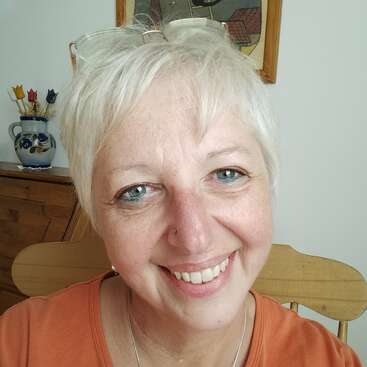 Eine lächelnde Frau mit kurzen weißen Haaren und blauen Augen sitzt auf einem Holzstuhl, trägt eine Brille auf dem Kopf und ein orangefarbenes Oberteil, drinnen mit Kunst.