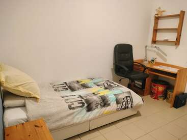 Dieses Bild zeigt ein einfaches, aufgeräumtes Schlafzimmer mit einem Einzelbett, einem Nachttisch aus Holz, einem Eckschreibtisch, einem Bürostuhl, einem Regal, einer Schreibtischlampe und einem gefliesten Boden. Minimalistische Einrichtung.