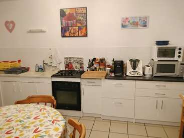 Diese gemütliche Küche verfügt über weiße Schränke, verschiedene Geräte, eine bunte Tischdecke, Holzstühle, Wandkunst, einen Geschirrständer, Utensilien und einen gefliesten Boden. Einladende Atmosphäre.