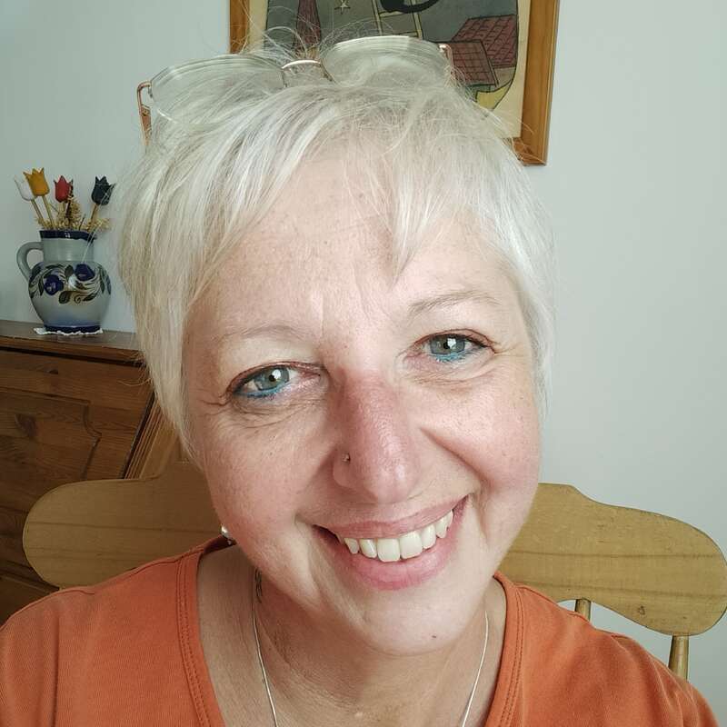 Eine lächelnde Frau mit kurzen weißen Haaren und blauen Augen sitzt auf einem Holzstuhl, trägt eine Brille auf dem Kopf und ein orangefarbenes Oberteil, drinnen mit Kunst.