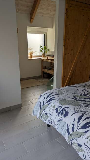 Un acogedor dormitorio con un edredón a flores, suelo de baldosas gris claro, puerta corredera de madera, vigas de madera a la vista y un pequeño rincón con plantas y una ventana.