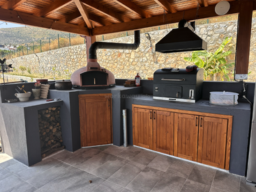 Cette cuisine extérieure comprend un four à pizza au feu de bois, un grill moderne, des armoires en bois, un espace de rangement pour le bois de chauffage et est abritée par un toit en bois, créant ainsi une atmosphère chaleureuse.