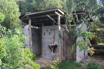 A imagem mostra uma pequena estrutura externa com um telhado inclinado, com uma fileira de janelas na frente, tendo como pano de fundo uma área gramada e uma cerca de madeira.