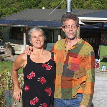 A imagem mostra um casal de meia-idade em pé do lado de fora de uma casa, com a mulher à esquerda usando um vestido preto e o homem à direita usando uma camisa multicolorida.