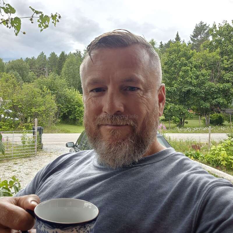 Un homme barbu aux cheveux courts sourit en tenant une tasse en plein air. De la verdure luxuriante, des arbres et une voiture garée constituent l'arrière-plan sous un ciel nuageux. Moment paisible.