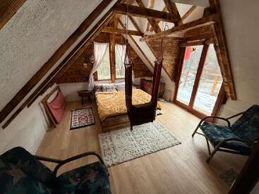 Una acogedora habitación en el ático con vigas de madera, ventanas grandes, una cama cómoda, dos sillas, una hamaca, alfombras y un balcón con unas preciosas vistas al exterior.