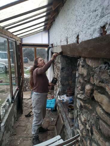 Una mujer está pintando una viga de madera en el interior de una terraza acristalada rústica con paredes de piedra. Hay diversas herramientas y botes de pintura esparcidos por todas partes, y la luz del día se filtra a través del techo de cristal.