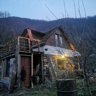 Ein rustikales, zweistöckiges Haus aus Stein und Holz steht in einer ländlichen, bergigen Gegend. Außenbeleuchtung und eine Flagge schmücken den Balkon, in der Nähe stehen Bäume.