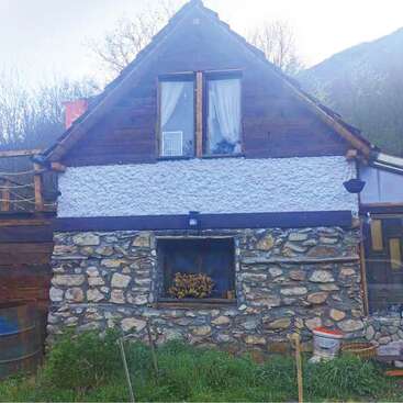 This image shows a rustic stone and wood house with a gabled roof, white textured walls, small window, greenery in front, and mountains in the background.
