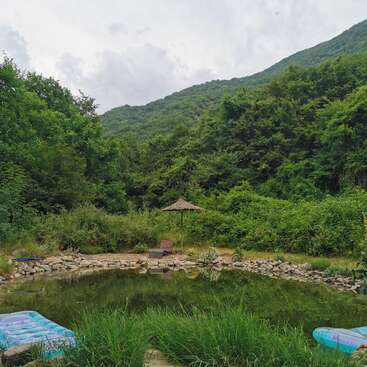 This image shows a tranquil, natural pond surrounded by lush green trees and hills. There are two blue mattresses and a sunbed with a parasol nearby.