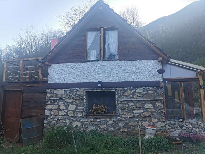 Una cabaña rústica de piedra y madera con una ventana abuhardillada, cortinas blancas y un pequeño balcón, rodeada de césped y árboles en un entorno rural y tranquilo.
