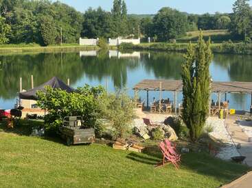 Un apacible refugio junto al lago con exuberante vegetación, asientos de madera en la cubierta, una tienda con toldo, muebles de exterior, árboles y un pequeño vehículo utilitario a plena luz del sol.