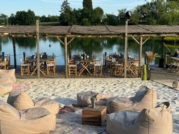 Un entorno sereno junto al lago con una zona de asientos de arena, pufs de color beige, mesas de madera, sillas bajo una pérgola, aguas tranquilas, vegetación y un ambiente relajado al aire libre.