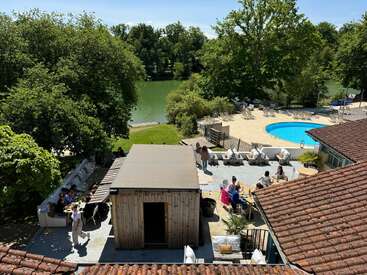 Esta imagen muestra una soleada zona al aire libre con gente socializando, una piscina, asientos en el patio, grandes árboles y un pintoresco río al fondo rodeado de vegetación.