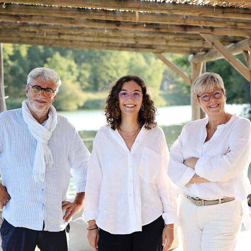 Tres personas, dos adultos mayores y una mujer más joven, sonríen bajo una pérgola de madera junto a un lago. Todos llevan ropa clara y gafas, y parecen felices.