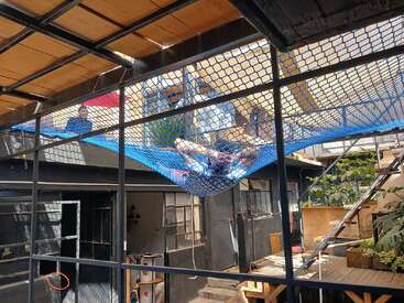 Une personne se détend sur un filet bleu suspendu au-dessus d'une cour. Il y a des plantes, des meubles en bois et un homme qui se tient à proximité. Le décor est créatif et urbain.