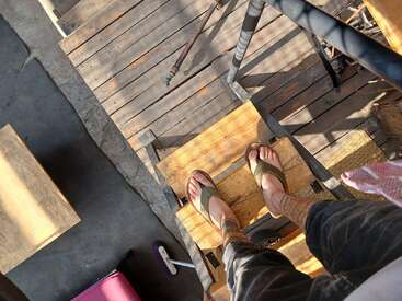 Une personne portant des sandales vertes et des chaussettes à motifs se tient debout sur un escalier en bois. La lumière du soleil projette des ombres. Des planches de bois, un balai et un objet rose sont visibles à proximité.