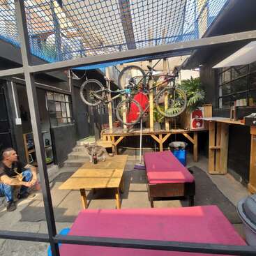 Ein gemütlicher Hof im Freien mit rosa Bänken, Holztischen und Fahrrädern, die an einer Wand hängen. Eine Katze sitzt auf dem Tisch, und ein Mann sitzt in der Nähe.