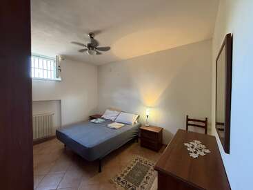 Se trata de un dormitorio sencillo con cama de matrimonio, mesilla de noche, lámpara, cómoda de madera, silla, ventilador de techo, ventana pequeña, suelo de baldosas y decoración minimalista.