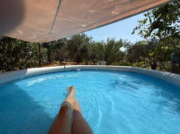 Una persona se relaja en una piscina redonda sobre el suelo bajo un toldo de sombra, rodeada de frondosos árboles y vegetación, disfrutando de un apacible día soleado al aire libre.