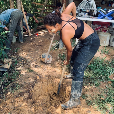 Une femme portant un pantalon noir et des bottes creuse un trou à l'aide d'une pelle, entourée de personnes travaillant sur un projet à l'arrière-plan.