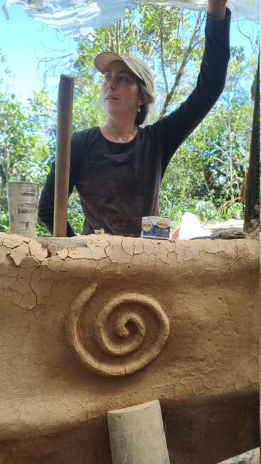 Una mujer con gorra está de pie detrás de un rústico muro de arcilla con un diseño en espiral, rodeada de árboles y herramientas, trabajando bajo una lona traslúcida a la luz del sol.