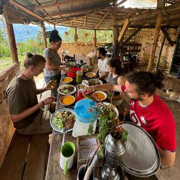 Cinq personnes partagent un repas à une table rustique en plein air sous un toit en bois, entourées par la nature, avec des plats colorés et une conversation amicale.