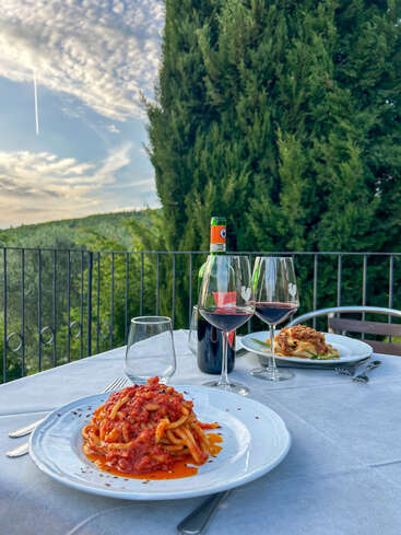 Das Bild zeigt ein ruhiges Abendessen im Freien mit einem Tisch mit weißer Tischdecke, einem Teller Pasta und zwei Gläsern Rotwein vor einer malerischen Kulisse aus Bäumen und blauem Himmel.