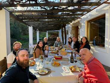 Un grupo de personas se sienta alrededor de una larga mesa al aire libre, disfrutando de una comida juntos a la luz del sol, con aspecto feliz y relajado, rodeados de naturaleza.