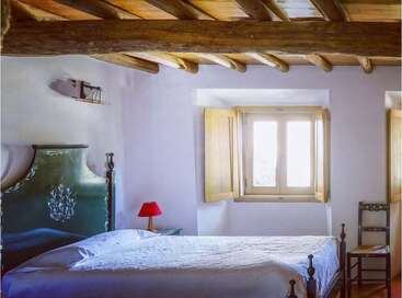Este acogedor dormitorio rústico cuenta con vigas de madera a la vista, un cabecero ornamentado en verde, una colcha blanca, sillas de madera, una lámpara roja y luz natural procedente de ventanas con postigos.