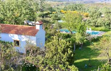 Una casa blanca con tejado de tejas rojas se encuentra entre frondosos árboles y jardines. Al fondo se ven una piscina y tumbonas.