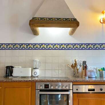Esta cocina presenta un salpicadero de azulejos con un borde decorativo, armarios de madera, electrodomésticos modernos, utensilios y una campana extractora de madera rústica bajo una cálida iluminación ambiental.
