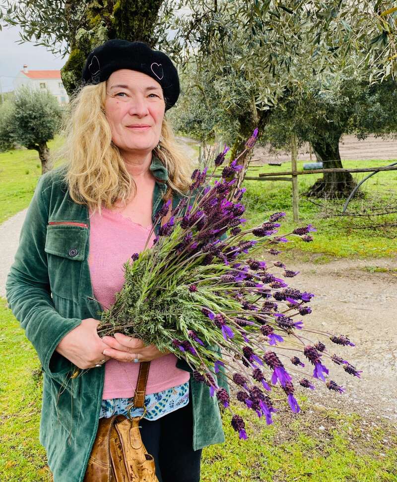 A woman with blonde hair, wearing a black beret and green jacket, stands outdoors holding a large bouquet of fresh purple flowers, surrounded by trees.